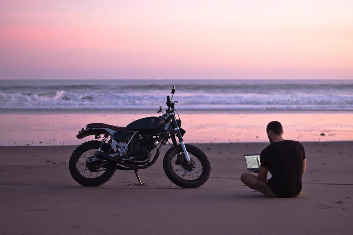 Lukas Rissing sitzt neben seinem Motorrad am Strand mit dem Laptop auf dem Schoß.